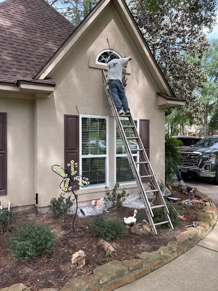 A person on an extension ladder works on a decorative arched window on the exterior of a tan stucco house.