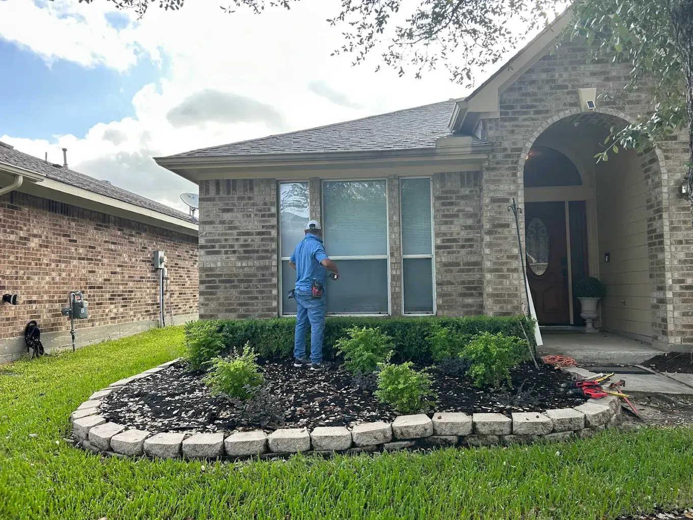 A worker in a blue shirt cleans a front window of a tan brick house with a landscaped garden bed in front.