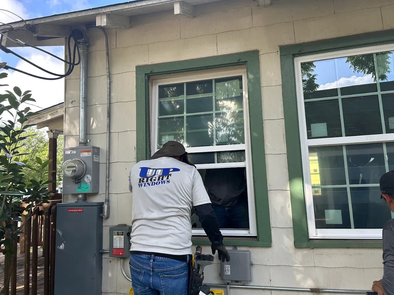 A worker in a light-colored uniform is installing or adjusting an open window on the side of a light-colored building.