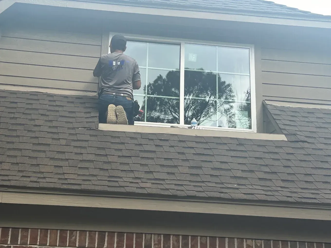 A worker sits on a sloped shingled roof, installing or cleaning a window on the side of a house.