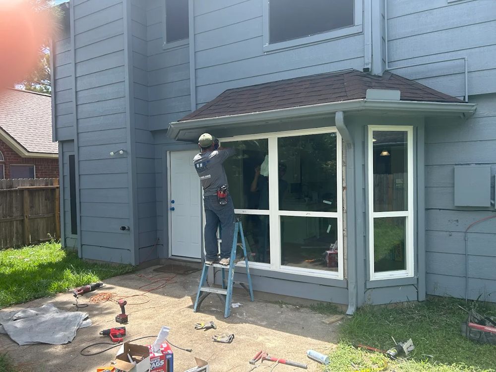 A person on a ladder installs a window into a blue, two-story house.