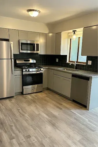 A modern kitchen with light gray cabinetry, stainless steel appliances, a black tiled backsplash, and wood-look flooring.