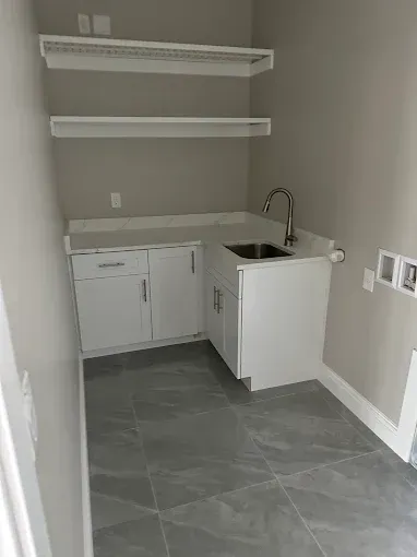 A small laundry room with grey tile floors, white cabinets, a sink, and two floating shelves against a grey wall.