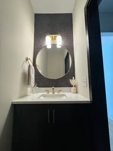 A modern bathroom vanity with a black cabinet, white countertop, round mirror, and patterned dark wallpaper behind it.