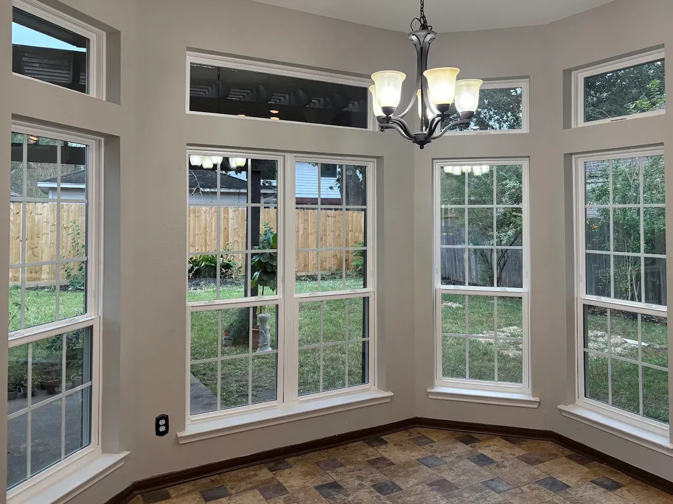 A sunny breakfast nook with large windows, tiled floors, and a hanging chandelier, overlooking a fenced backyard.