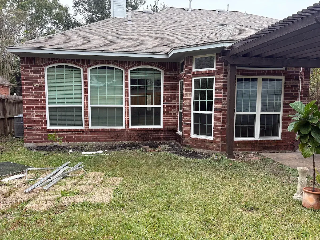 A backyard view of a one-story brick house with arched windows, a dark roof, and a wooden pergola on the right side.