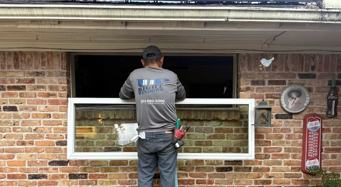 A person in a gray long-sleeved shirt installs a rectangular window frame into an opening on a red brick house wall.