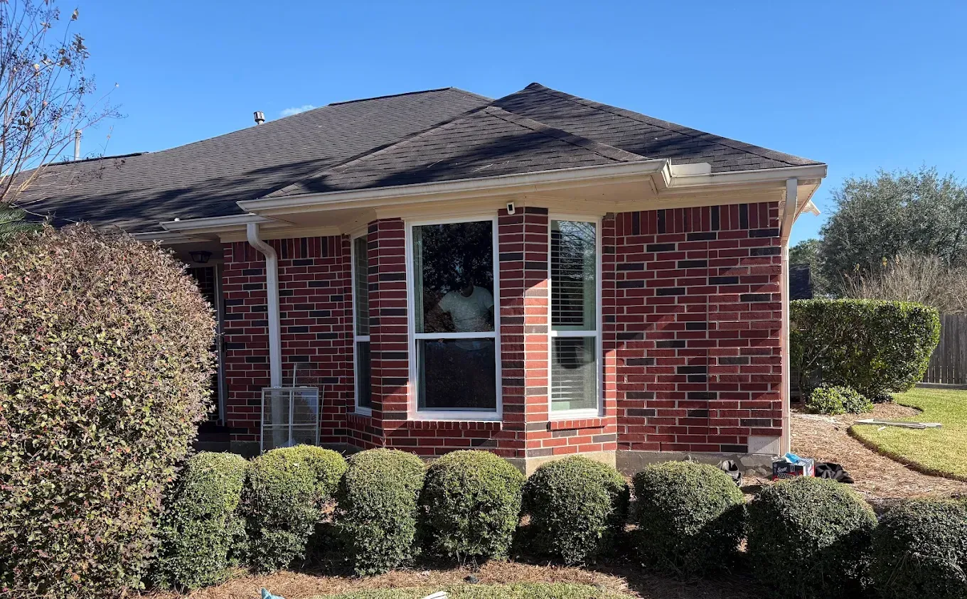 A single-story brick house with a dark shingled roof, windows, and a row of rounded bushes in the front yard.