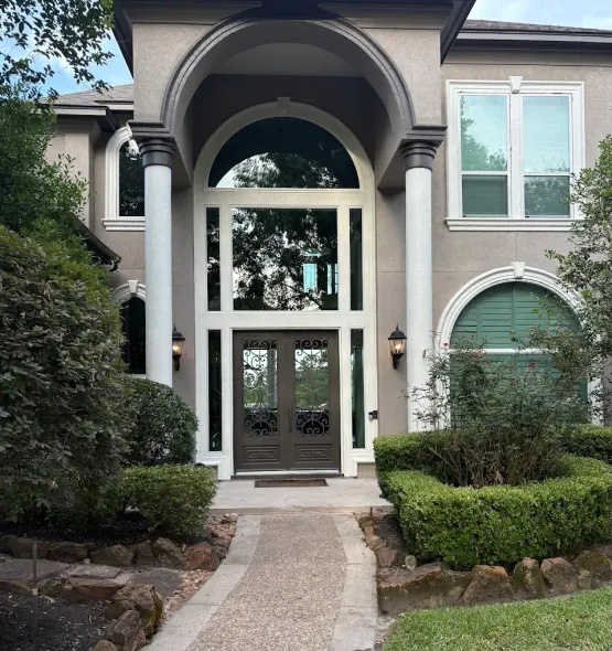 A two-story tan home with a covered front entrance featuring two white columns, arched windows, and brown double doors.