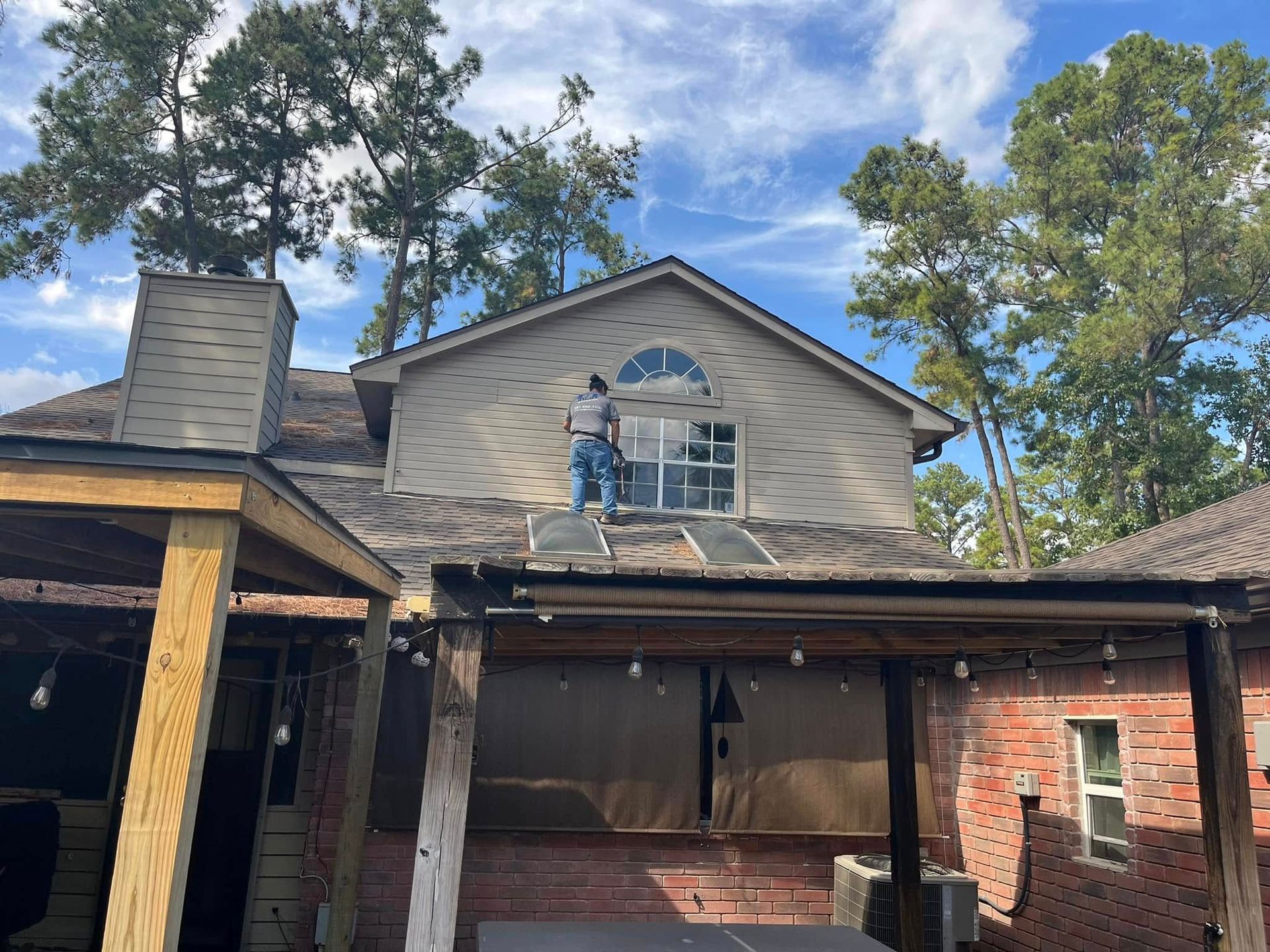 A person stands on a residential roof, near a second-story window, while performing work on the exterior of a home.