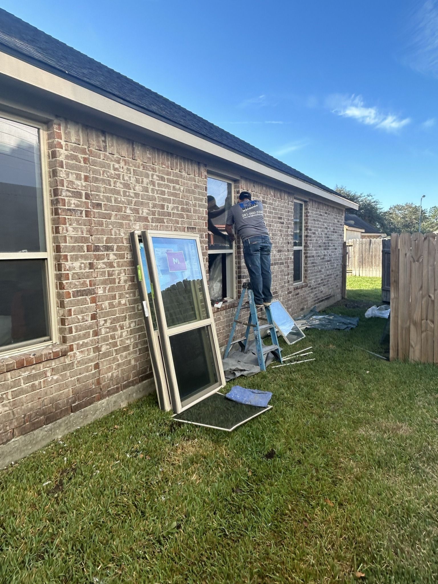 A worker on a ladder installs a window in the brick wall of a house exterior during a sunny day.