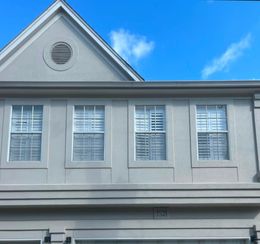 The exterior of a light gray two-story building featuring four windows and a triangular roof with a round vent.