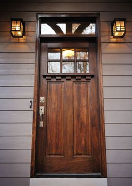 A dark wood Craftsman-style front door with a transom window, glass panes, and a silver handle, flanked by two lanterns.