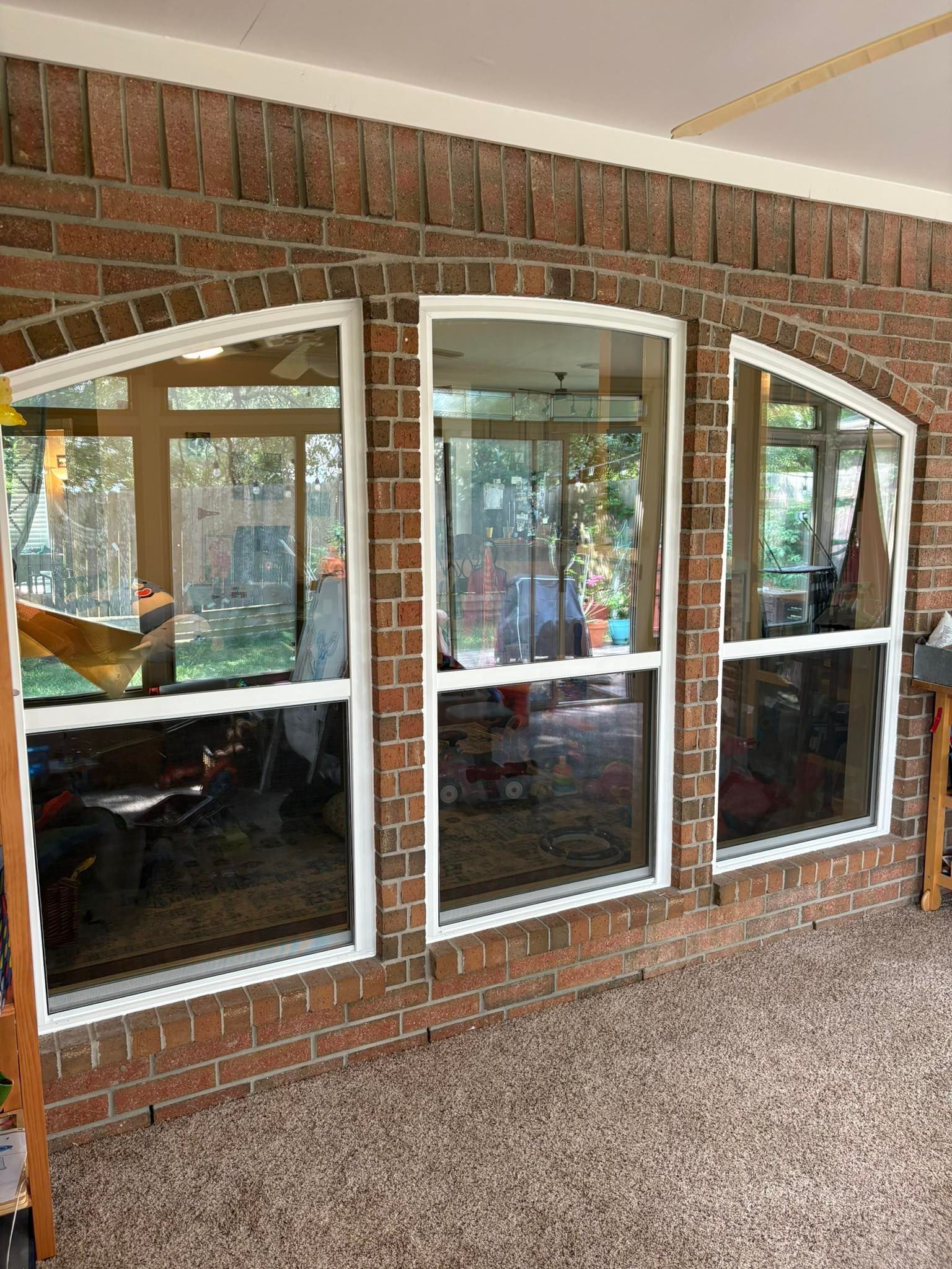 Three large, arched windows with white frames installed in a reddish-brown brick wall overlooking a sunroom.