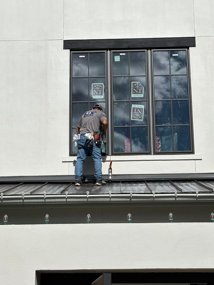 A person in a gray shirt and blue jeans works on a window installation on the exterior of a white building.