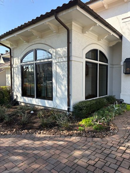 A corner view of a white stucco house with arched, black-framed windows, a dark roof, and a stone paver walkway.