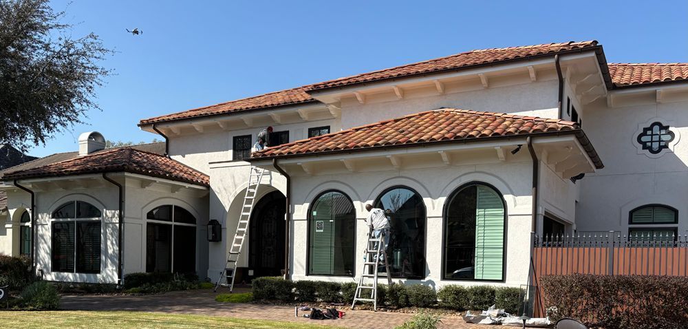 A painter stands on a ladder working on the exterior of a white, Mediterranean-style house with a tile roof.