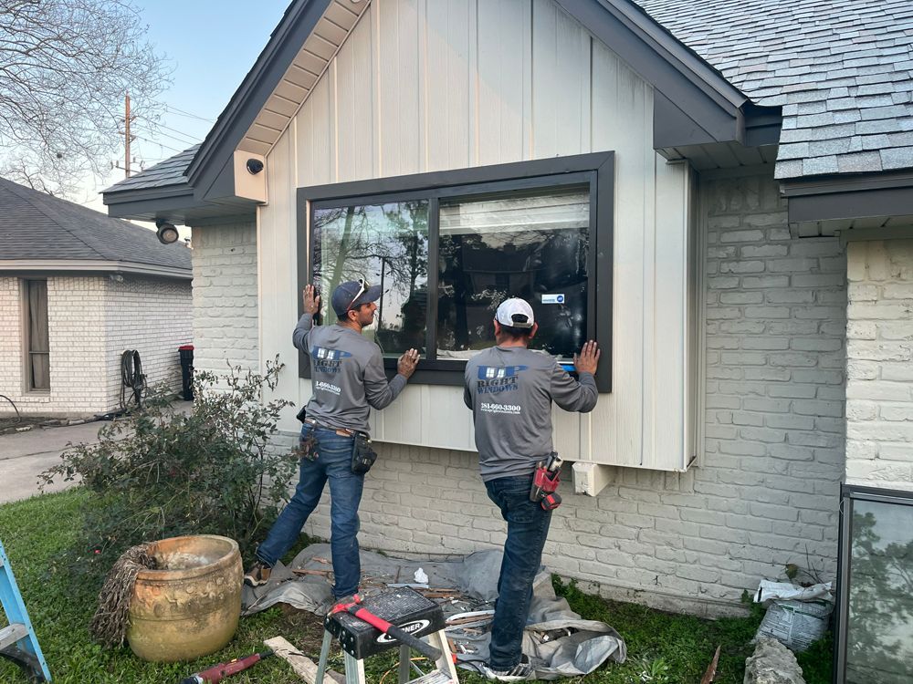 Two workers in grey shirts install a new dark-framed window on the front exterior of a light-colored brick house.