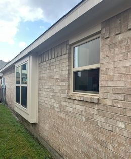A side view of a tan brick house with two windows and a tan trim, set against a lawn under a cloudy sky.