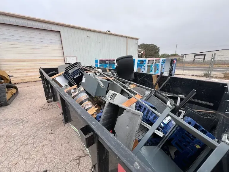 Black dumpster filled with scrap metal and office furniture, outside a warehouse.