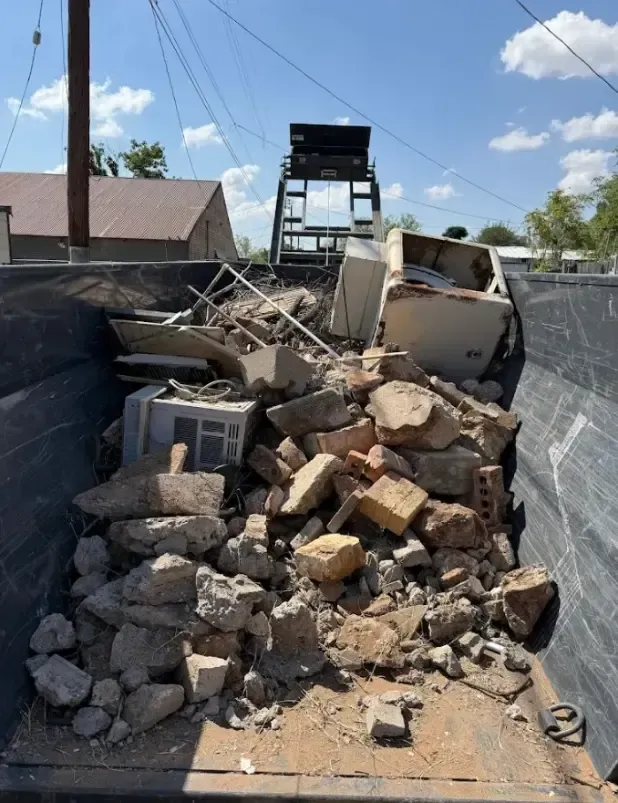 A dumpster filled with demolition debris, including bricks, concrete, and appliances, outdoors under a blue sky.