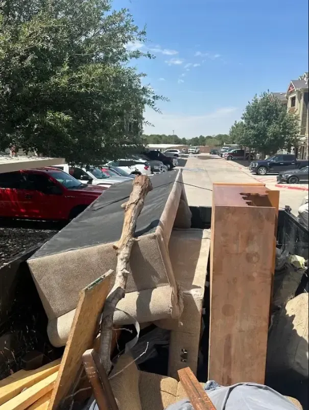 Dumpster overflowing with furniture and debris in an apartment complex parking lot under a blue sky.