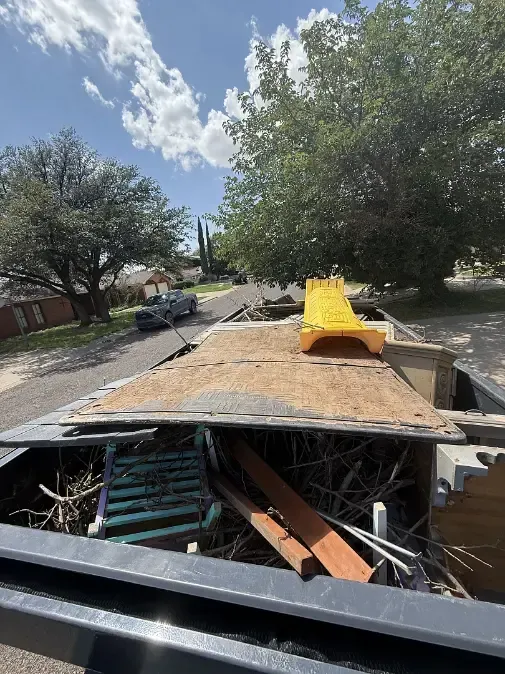 View from inside a truck bed filled with debris; yellow slide, wooden boards, tree branches visible. Street, houses, and sky background.