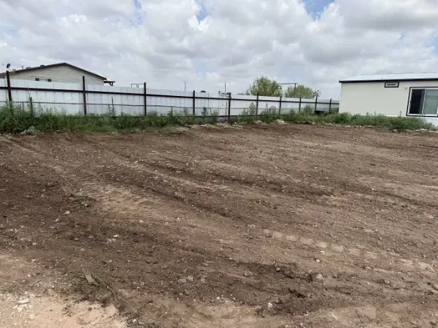 Dirt lot with tire tracks, chain-link fence, white building, and cloudy sky.