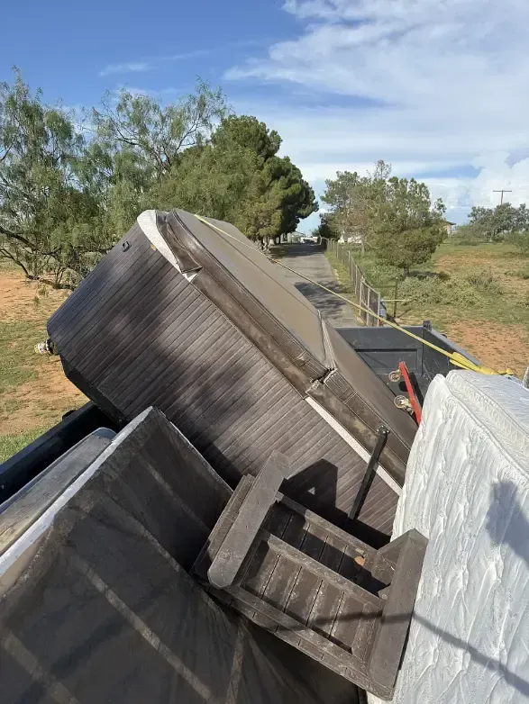 A truck bed filled with old mattresses and a hot tub. A rural road is in the background with trees.