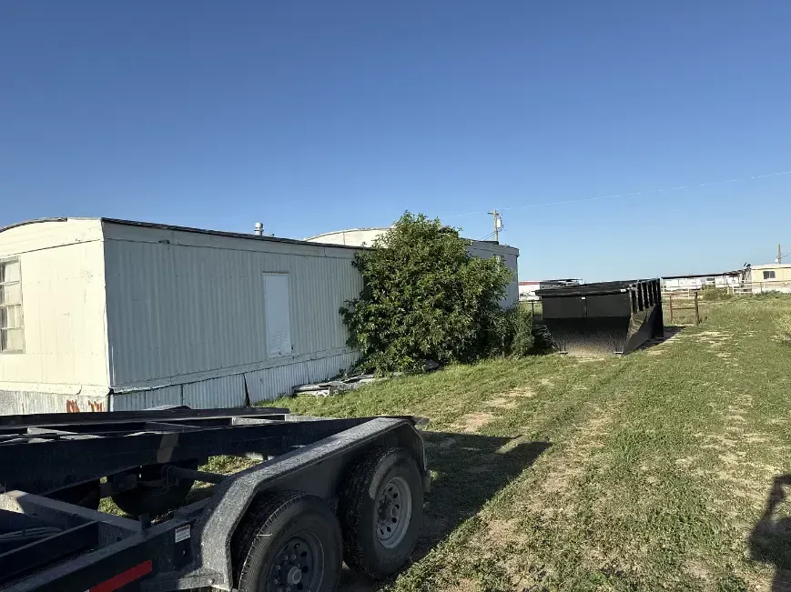A trailer near a mobile home with a dumpster in the background under a blue sky.