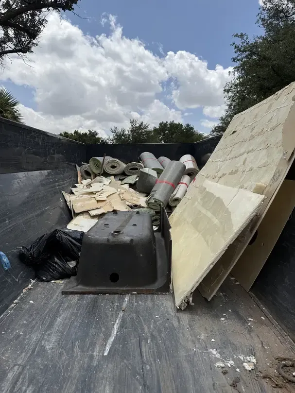 Dumpster filled with construction debris: rolls, panels, a sink, and garbage bags against a cloudy sky.