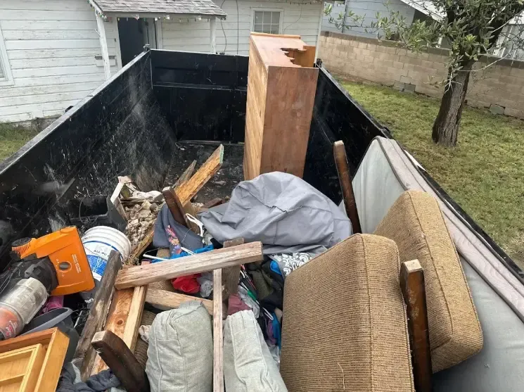 A dumpster filled with discarded furniture, wood, and debris outside a white building.