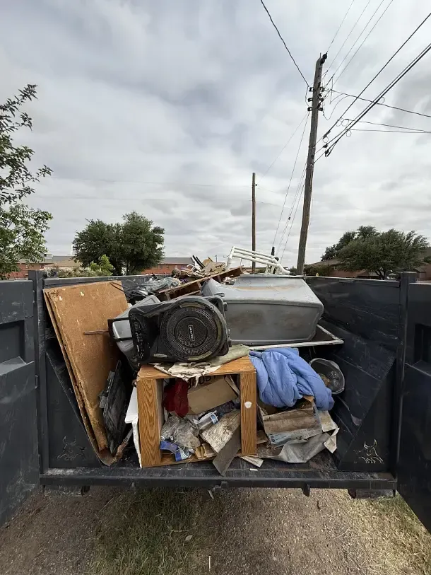 A truck bed filled with assorted discarded items, including a cabinet, board, and metal components. Cloudy sky backdrop.