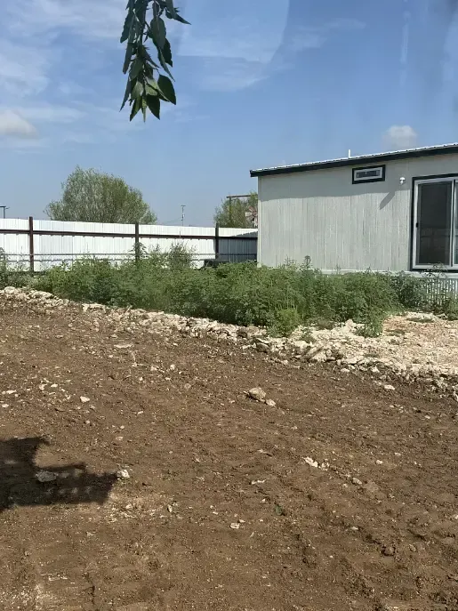 A newly tilled, brown dirt area in front of a white building with a grassy overgrown area. Blue sky.