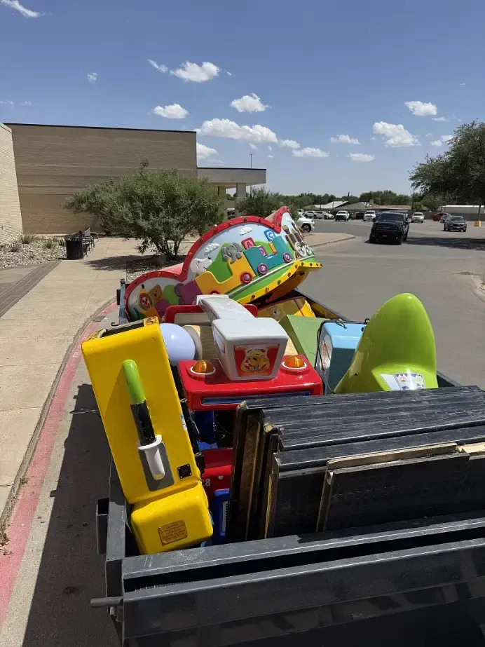 Pile of children's ride-on toys in a dumpster in front of a building on a sunny day.