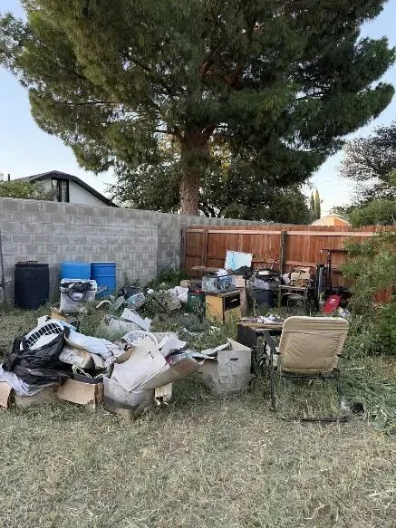A cluttered backyard with trash, old furniture, and a large tree against a wooden and concrete fence.