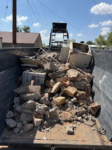 Dumpster filled with demolition debris, including concrete, bricks, and a small appliance, under a bright sky.