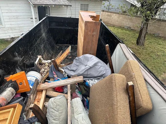 A dumpster filled with household waste, including furniture cushions, wood planks, and other debris.