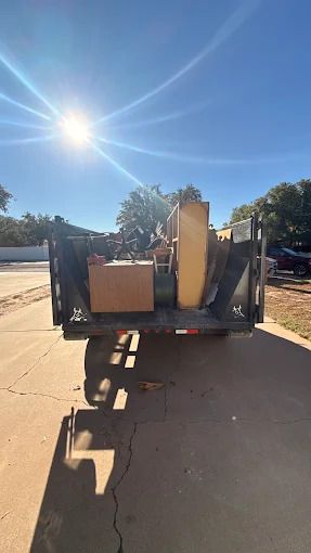 Dump truck bed filled with debris, parked on concrete. Sun shines brightly.