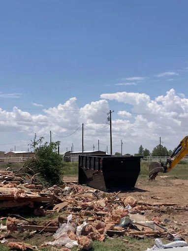 Demolition site with debris, a dumpster, and an excavator under a partly cloudy sky.