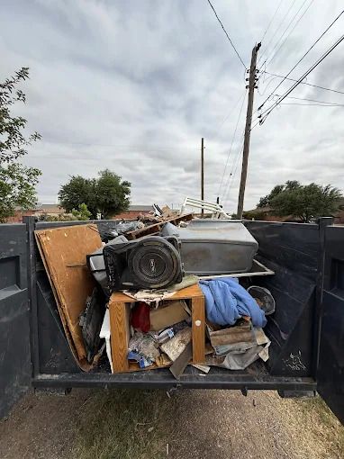 Dump truck bed filled with assorted trash, including wood, electronics, and debris; overcast sky in the background.