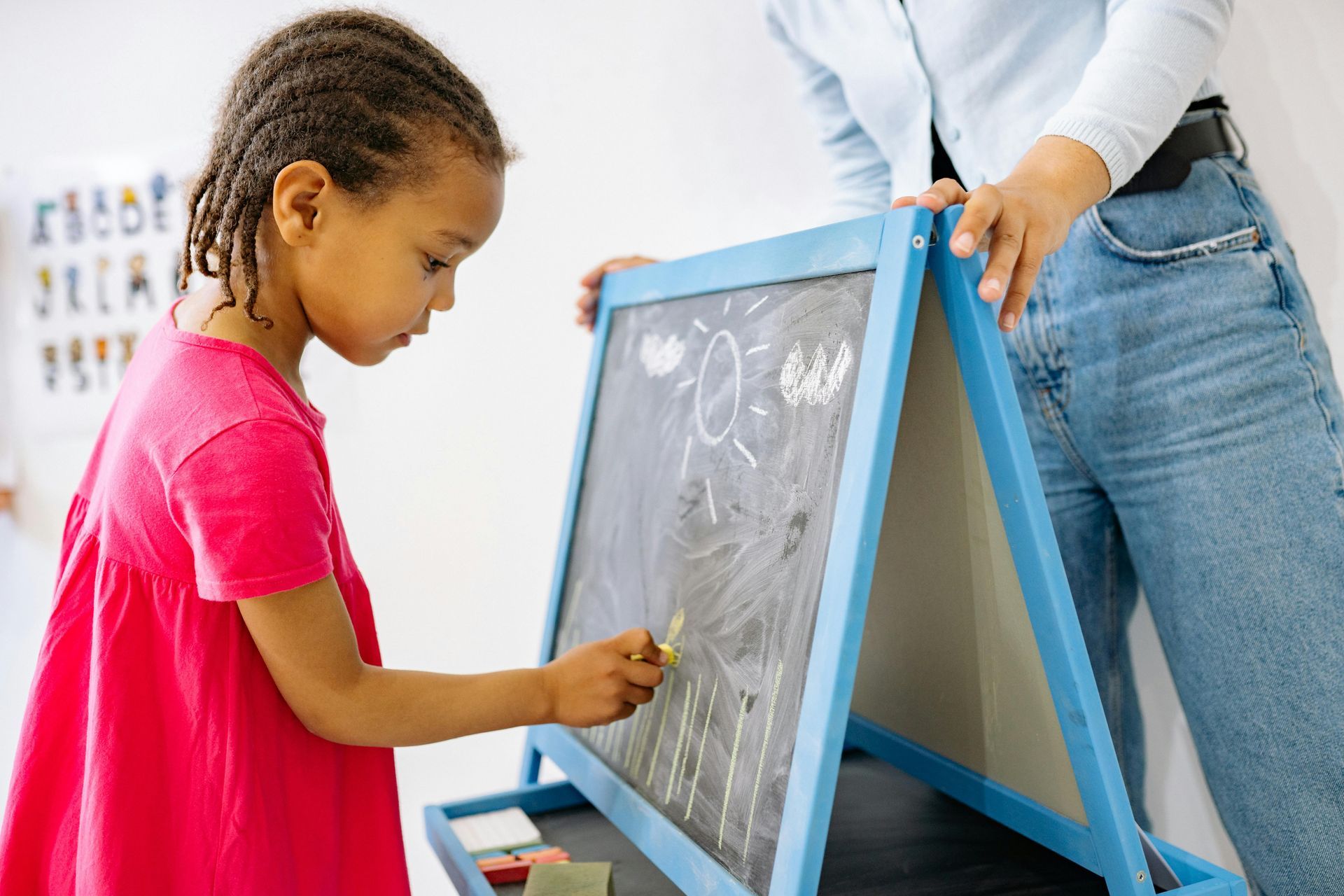 Girl in pink dress drawing on a chalkboard, with adult standing beside her.