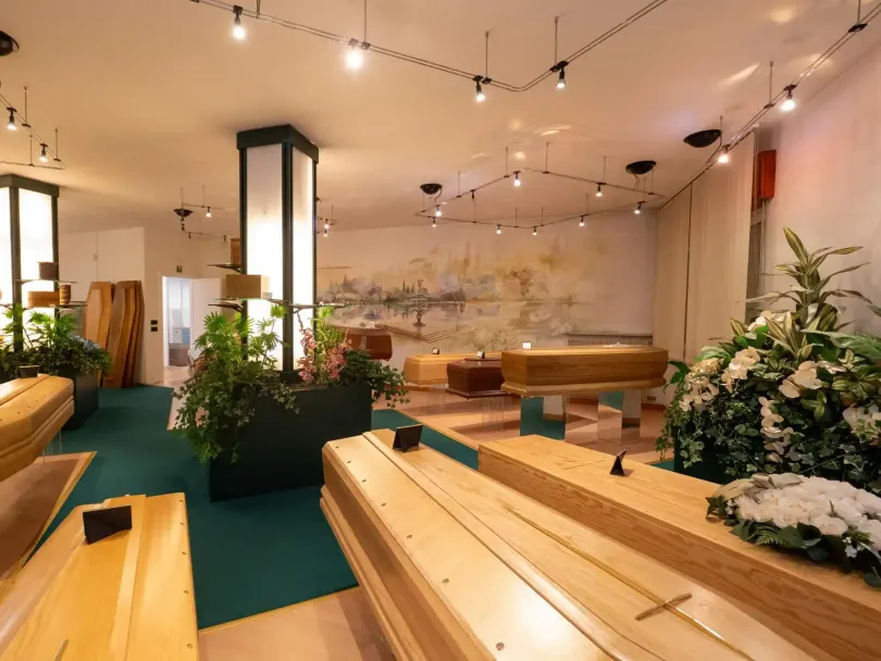 Interno di un'agenzia di pompe funebri con diverse bare di legno, piante e luci soffuse.