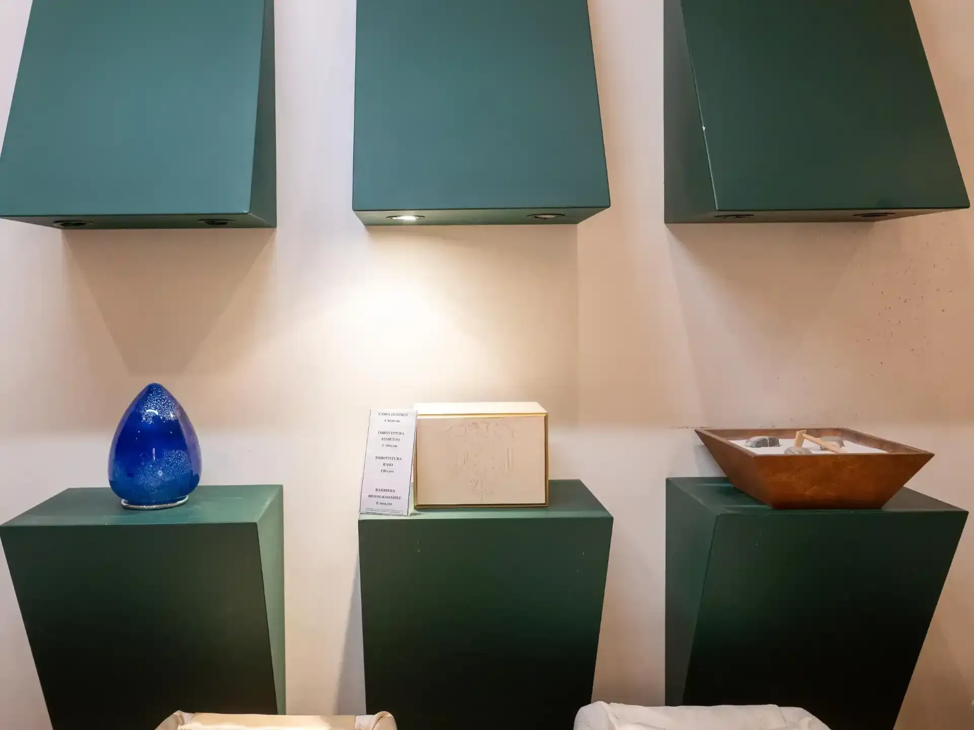 Espositore a tre livelli con piattaforme cubiche verdi che sostengono un vaso blu, un blocco color crema e una ciotola di legno, sotto una decorazione murale geometrica verde.