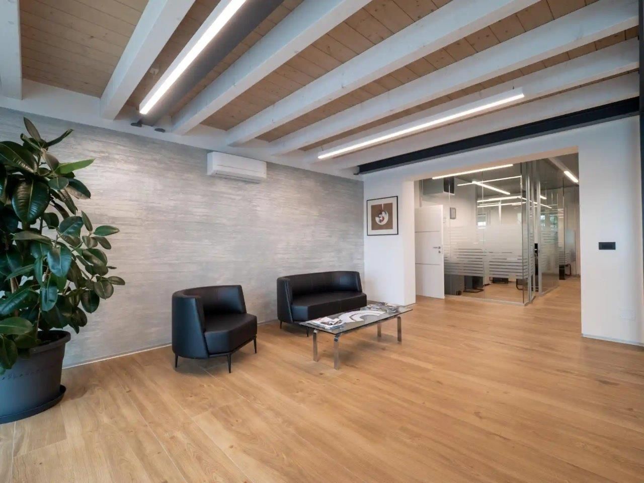 Moderna sala d'attesa con pavimenti in legno, soffitto con travi a vista, sedie in pelle nera e piante in vaso.