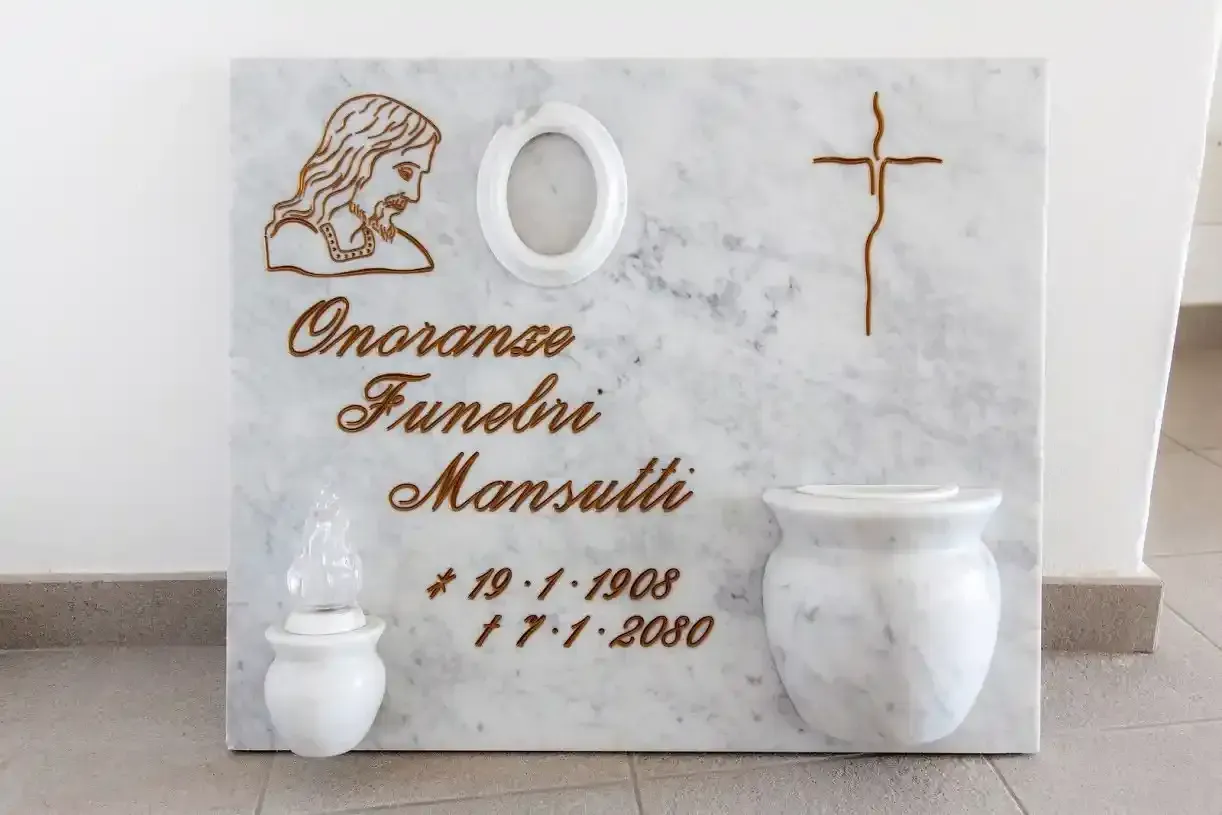 Lapide in marmo con testo inciso e simboli religiosi, tra cui un volto, una croce e un'urna.