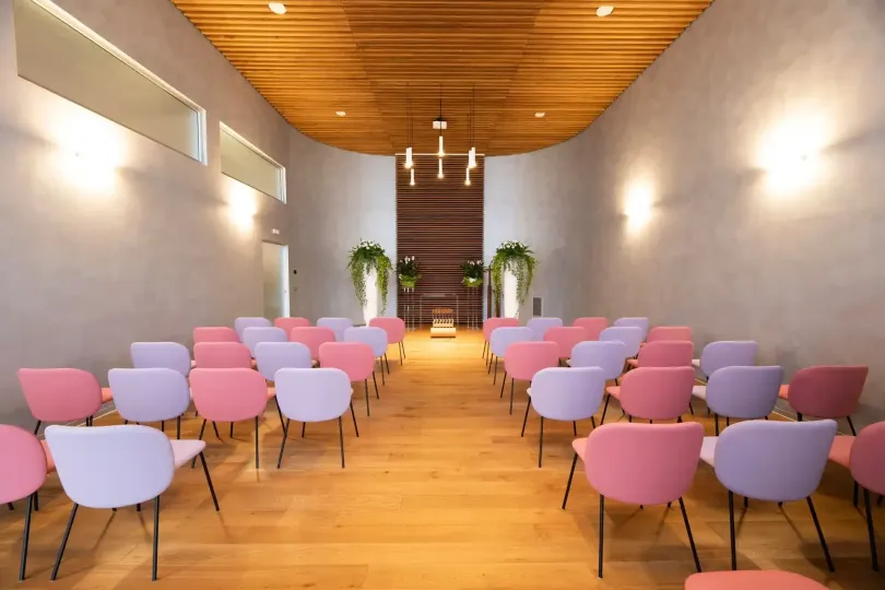 File di sedie rosa e viola in una stanza curva con pavimento e soffitto in legno, illuminata da applique.