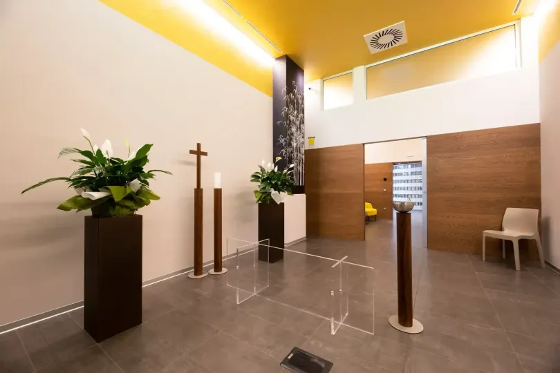 Corridoio con composizioni floreali, dettagli in legno e una croce. Soffitto giallo, pavimento in piastrelle grigie, pareti bianche.