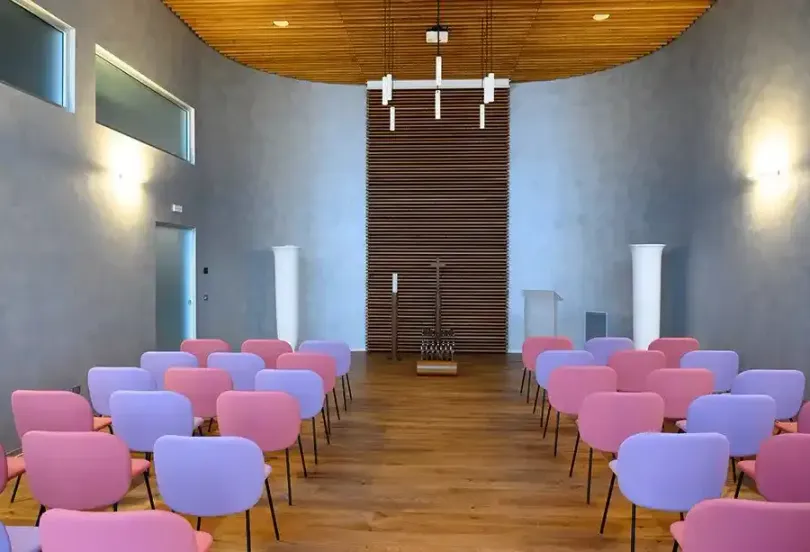 File di sedie rosa e viola in una cappella con pavimenti in legno e una parete posteriore in legno, illuminata da lampade.