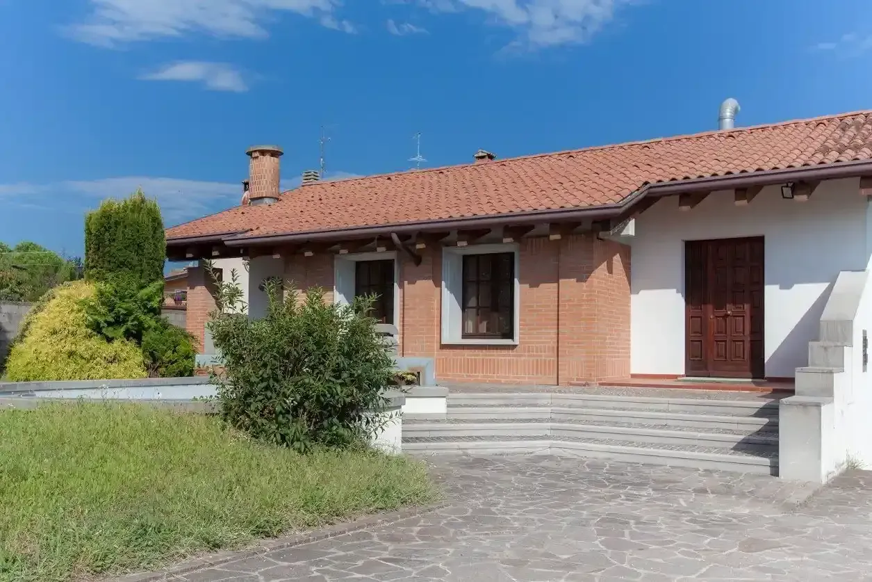 Una casa a un solo piano con tetto di tegole rosse, muri di mattoni e una porta di legno sotto un cielo azzurro brillante.